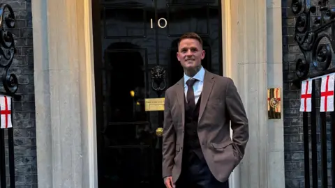 A smiling man stood outside the front door of Number 10 Downing Street. He is wearing a brown suit jacket with black waistcoat underneath, white shirt and brown tie. Attached to some black railings are St George's flags.