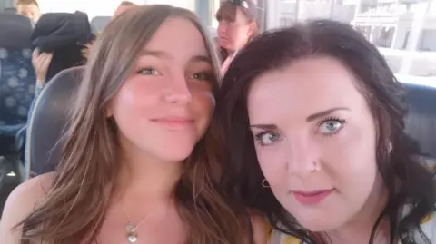 Chloe Longster (left) smiles with her mother Louise Longster (right). Chloe has long brown hair that has been curled, she is wearing natural makeup and has a silver locket around her neck. Her mother is looking at the camera and has dark brown hair, light red lipstick and a pierced nose. They are sitting on a coach together.