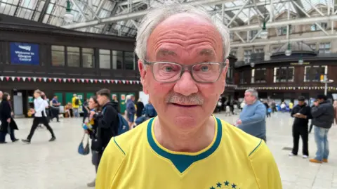 Malcolm McLean, an older man wearing glasses and a bright yellow Brazil national football jersey stands in the foreground of a large indoor train station concourse. He is facing the camera. The spacious hall features a high glass-and-steel roof, dark wooden buildings along the sides, and people walking in the background. A sign reading “Welcome to Glasgow Central” is visible behind him.