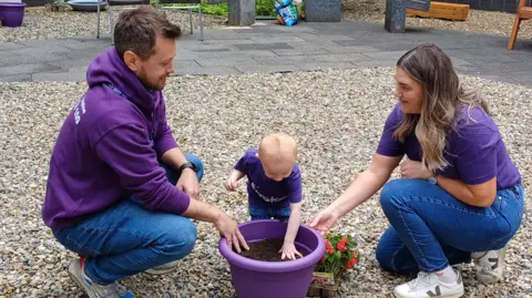 Stroke Association Shows a man on the left, children in the middle and woman on the right, all wearing purple tops and playing with a flower pot. 