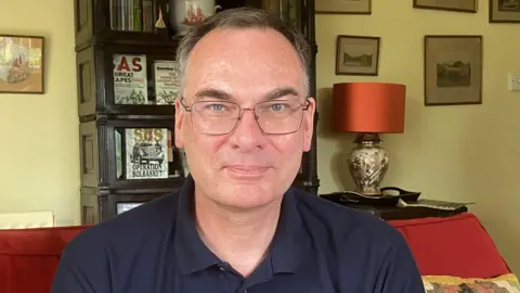 Dr Will Ward looking at the camera, has blue eyes, grey hair and is wearing glasses and a navy polo shirt. In the background is a living room with a bookcase.