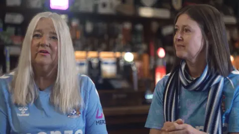 Kieran Doherty A female Coventry City fan wearing a sky blue Coventry City top is sitting on the left with blonde hair looking to the camera. Another female Coventry City fan wearing a sky blue Covenrty City top sits on the left also wearing a dark blue, sky blue and white scarf. She has brown hair and is looking at the blonde-haired woman.