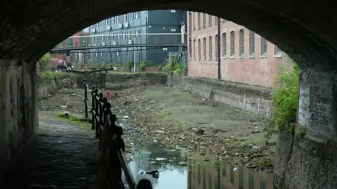 A canal bridge over a canal with almost all of its water drained out. Rubbish is left amid a pool of water left.
