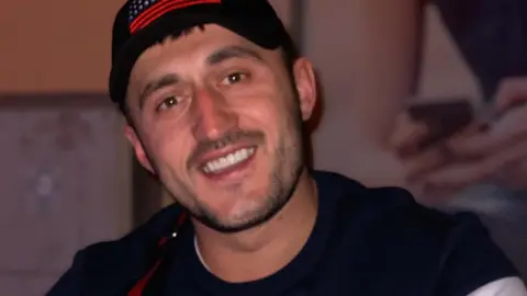 Sali Byberi is smiling at the camera and is wearing a black top and a black cap. He has black stubble.