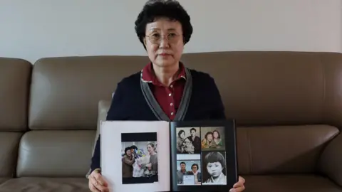 BBC Korean Han Tae-soon looks at the camera while sitting on a brown leather sofa, holding a photo book filled with pictures of her daughter Kyung-ha