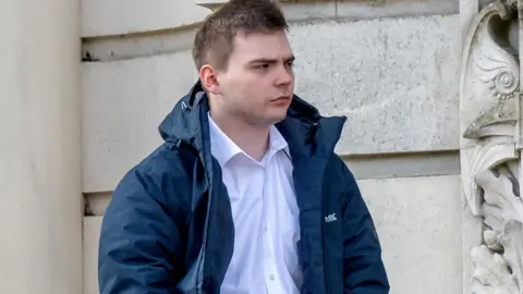 Oliver Beynon stands outside court in a white collared shirt and dark blue jacket, looking off to the side with a serious expression.