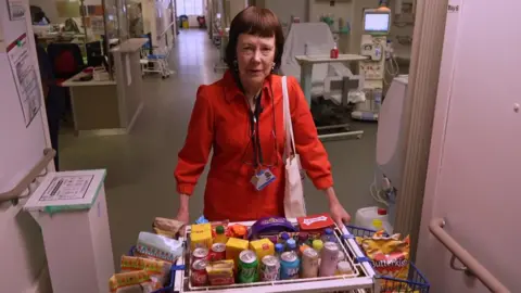 Honor standing in a hospital ward in a red coat. She has both hands placed on a trolley full of refreshments.