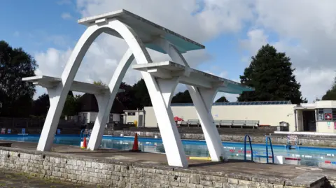The outdoor lido pictured on a sunny day. There is a tall arched diving board in the forefront, sitting on top of a low brick wall. Behind it is the filled pool, with blue, white and red plastic floats separating the swimming lanes and a ladder descending into the water.