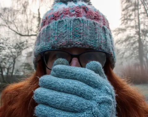 Ian Hawley Close-up picture of a woman wearing blue woollen gloves. The woman, who has long red hair and a blue woollen hat on, is holding her hands over her face, as if trying to warm them with her breath. She is wearing glasses and is looking directly at the camera.