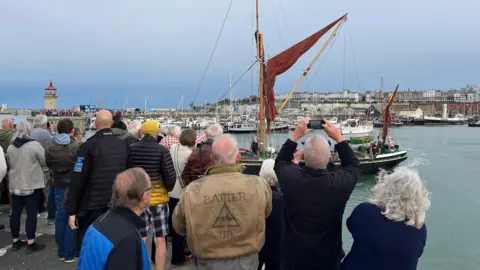 People standing by a harbour and watching a boat set off.
