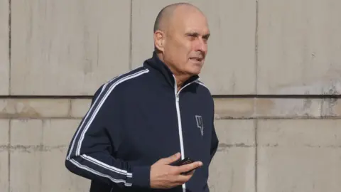 Derek Lammey, a man with a shaved head, pictured outside court at a previous hearing. He is wearing a navy sports jacket with white stripes on the sleeves and is holding a mobile phone.