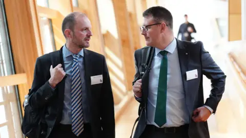 PA Media Stephen Gethins wearing a dark suit, blue shirt and striped tie. He is hold a rucksack over his shoulder. He is walking down a corridor in Holyrood next to David Linden, also wearing a suit with a green tie.