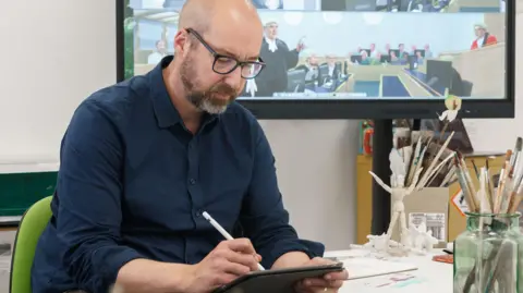 University of Sunderland Nick Lewis sketches on his iPad using a white stylus pencil. He is wearing a navy shirt and glasses and is bald with a dark and grey beard. Behind him is a TV screen showing one of his sketches form the trial, which depicts a barrister standing and speaking to a judge.