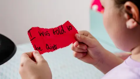 Futuros Valientes Una niña, cuyo rostro solo se ve de perfil, mira un trozo de papel rojo que sostiene con ambas manos. En el papel, escrito con letra infantil y tinta negra, se lee: «Me dijeron que guardara silencio».  
