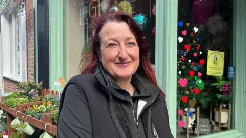 Ellen Knight/BBC Jenny McHale is outside her shop. She is wearing a black fleece with a black gilet over the top, and has long red hair. The shopfront is painted light green, with trays of flowers and plants arranged in rows outside. The shop windows are decorated with heart stickers. 