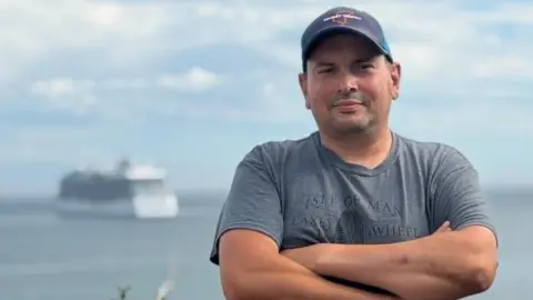 Frank Schuengel Frank has his arms folded, is wearing a grey t-shirt and TT cap. Behind is the sea with a large cruise ship blurry on the left.