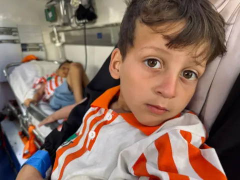 A boy in a red and white shirt sits on the ambulance, with another in a similar shirt lying down behind