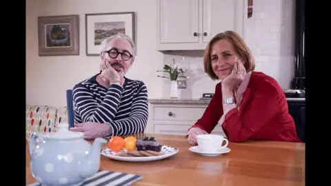 Alan Cumming and Kirsty Wark sit at a kitchen table with cups of tea and a plate of biscuits in front of them