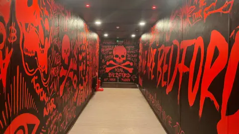 Tony Fisher/BBC The players' tunnel at Real Bedford's football ground, with black walls and red lettering with a skull and crossbones at the end, and the words "Real Bedford".