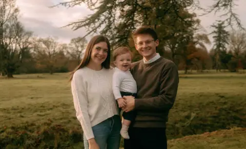 Becy/Bell Art Photography Ashlyn and Joe stand in front of a green field backdrop with trees, as they hold a baby in their arms while Ashlyn wears a cream jumper and Joe a brown jumper.