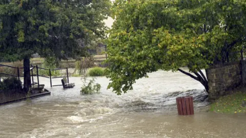 Flooding in Calne with trees and structures partially submerged in grey flood water, which is swirling around