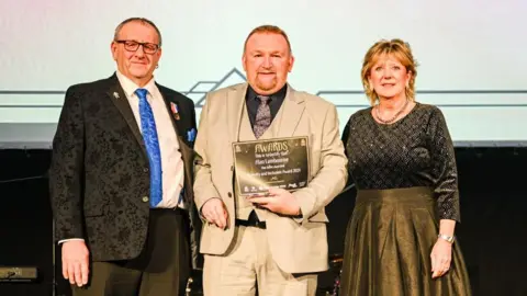 A man in a beige suit and tie stands in between a woman wearing a green and golden dress and another man dressed in a black suit and blue tie. The man in the beige suit is holding an award certificate.