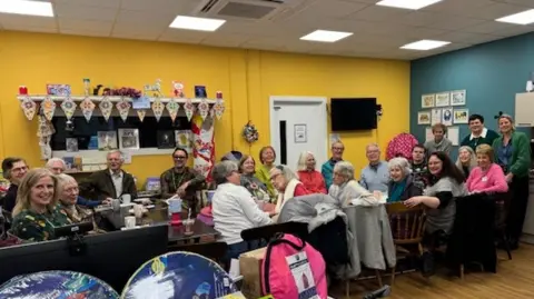 A long table with multiple people sat along it. They have turned to look at the camera and are smiling. The room has colourful walls decorated with crafts.