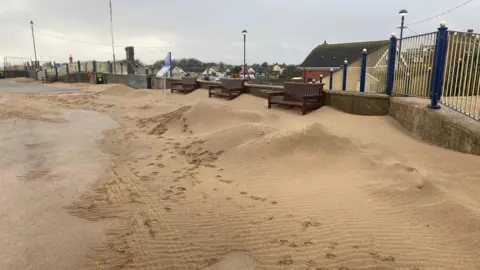 The photo shows a beach walkway. To the right hand side are piles of sand, which are almost as high as the wall in the background which is running adjacent to the path. There are three benches just in front of the wall. They are blocked off by the piles of sand. 