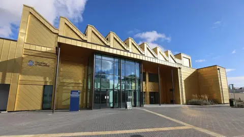 A multi-storey building with a shiny gold exterior and a large glass entrance is photographed on a sunny and largely clear day. 