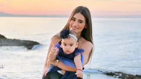 A woman with long dark hair smiles as she holds a toddler in a dress on a beach, the sunset behind her