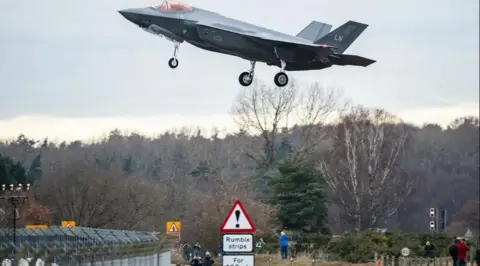 Getty Images A United States fighter aircraft with a twin tail coming in to land at an RAF base. The perimeter fence can been seen as well as civilian aircraft spotters. There is a road sign in the foreground