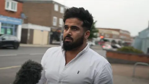 BBC Jamal is wearing a white t-shirt and has a microphone pointed towards him 