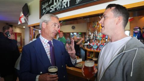 Nigel Farage and James McMurdock stand a bar holding pints while speaking to one another. 