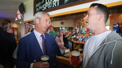 PA Nigel Farage and James McMurdock stand a bar holding pints while speaking to one another. 