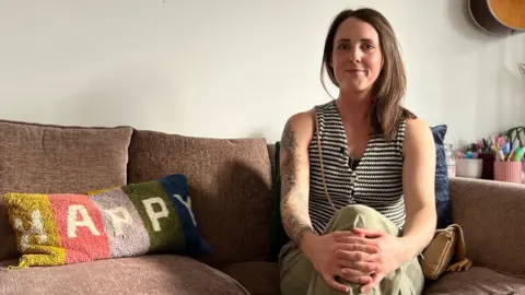 BBC Sammy Kean sits on a brown sofa with her legs crossed and her hands holding her knee. She is wearing a striped vest and pale green trousers. Next to here is a cushion that says 'happy'.