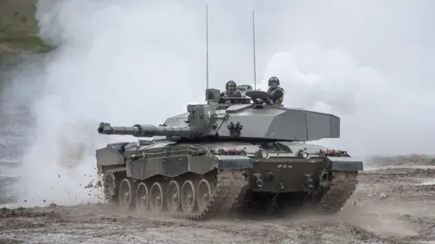 PA Media A tank pictured in Wiltshire in 2017. Two members of the armed forces drive through mud, with smoke in the background.