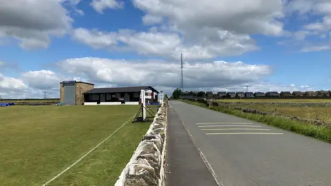 The top of a drystone wall with a cricket pitch on the left bordered by a road and a field on the right with a row of houses in the distance.