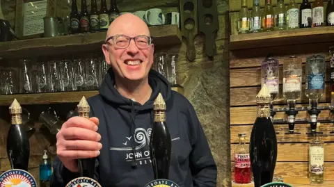 John has a hand on a beer engine handle in the brewery's bar as if about to pull a pint of beer. There are rows of bottles and glasses on the wall behind him.