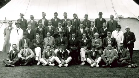 Essex County Cricket Club A black and white team photo taken in front of a large white tent. The male players are either sitting or standing across four rows. They are wearing smart outfits.