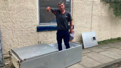 A man in a grey top stands in a fly-tipped fridge pointing at it.