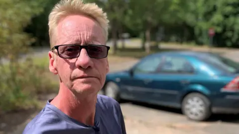 A man wearing sunglasses looks into the camera. A car can be seen in the background.