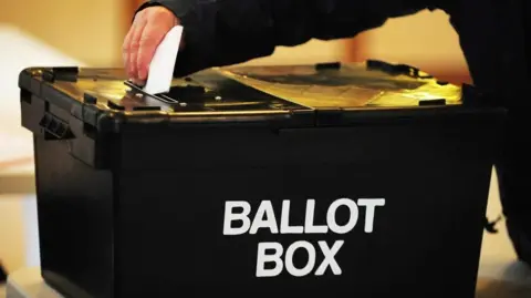 BBC A stock image showing a hand putting a folded ballot paper into a black plastic box with ballot box written on the side in white lettering