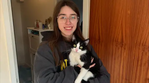 Abi Left A young woman holding a black and white cat in front of a wooden door. The woman is wearing a grey hoodie and clear-framed glasses. 