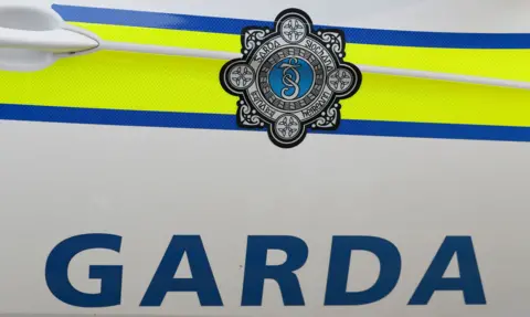 Close up of Garda emblem on patrol vehicle door. Shows logo on a bright yellow background, with blue lettering below reading 'GARDA'.