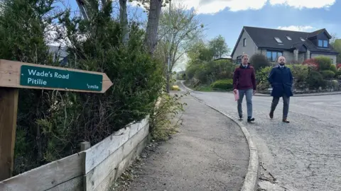 Two police detectives walk down a road with a sign saying Wade's Road, Pitilie in the foreground