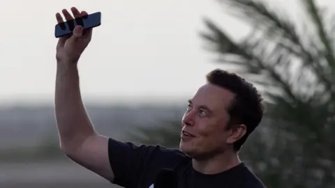 Elon Musk wearing a black T-shirt and holding his phone above his head.