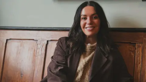 Gwen Matthews A woman with brown hair, in a brown jacket, sits on a wooden bench facing the camera.