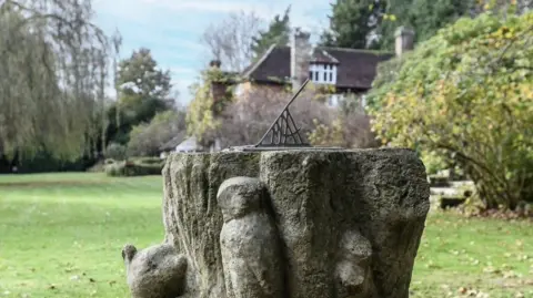 Ashdown Forest Foundation A weathered stone garden feature with a metal sundial set on top, engraved with numbers, stands in a lawn at Cotchford Farm, with shrubs, trees and the farmhouse visible in the background.
