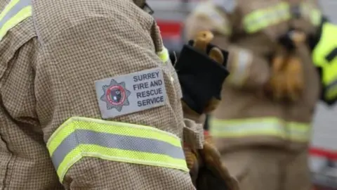a close-up of the arm of a fire fighter at a scene, displaying the sleeve badge with Surrey Fire and Rescue Service and its logo stitched on it and a fluorescent yellow and grey striped band around the sleeve