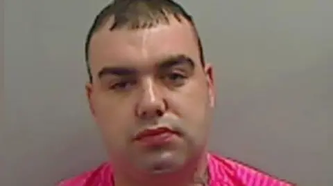 A mugshot of John McElvaney. He has short dark hair and is wearing a pink T-shirt. A curling tattoo can be seen on his neck.